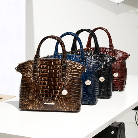 Four structured Brahmin handbags displayed together in brown, deep blue, black, and red embossed leather. Each bag features a glossy crocodile texture, rounded double top handles, a spacious trapezoid silhouette, gold tone hardware, and a hanging Brahmin medallion charm.
